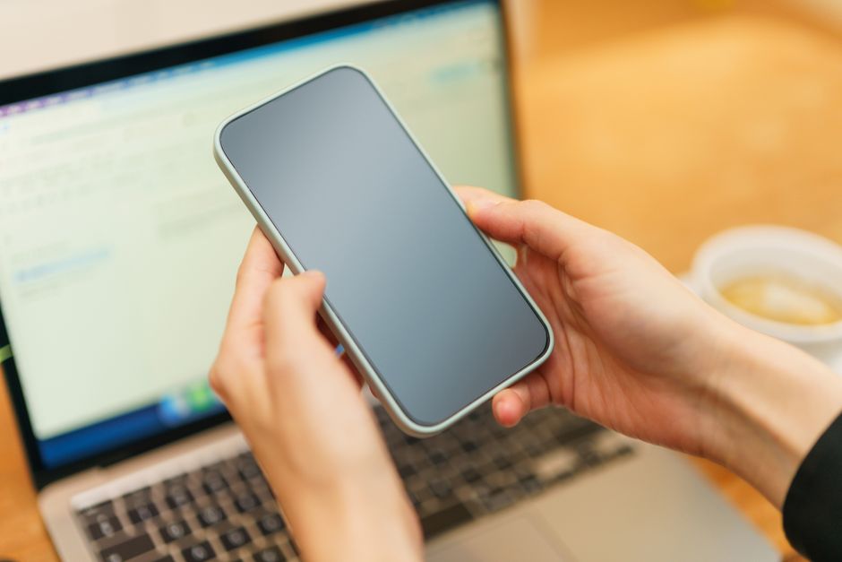 Person holding a smartphone above an open laptop with coffee in the background, ready for work or communication.